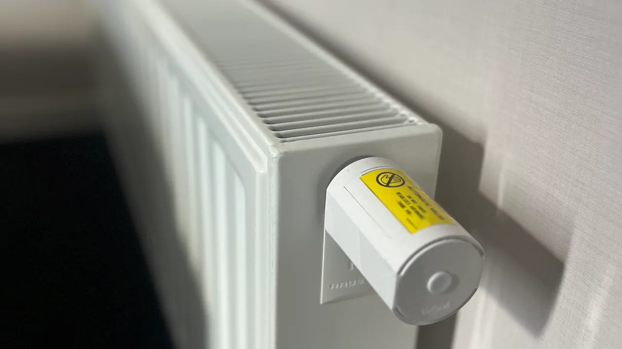 Een witte radiator gemonteerd tegen een lichtgekleurde muur, voorzien van een slimme thermostaatknop met een gele waarschuwingssticker. De thermostaat heeft een modern ontwerp en biedt geavanceerde temperatuurregeling. Zonlicht werpt een schaduw op de muur, wat zorgt voor een warme en huiselijke sfeer.