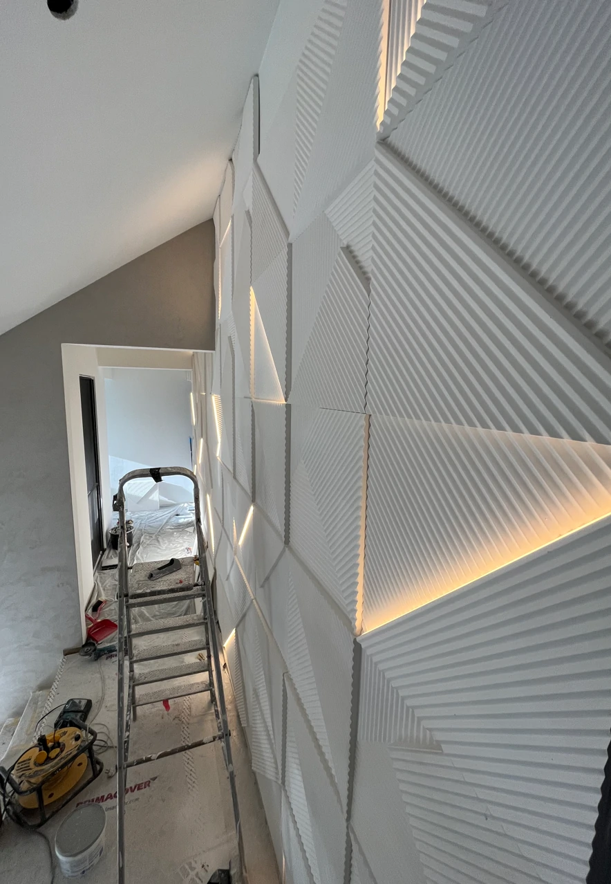 Een modern interieur met een decoratieve wandbekleding bestaande uit witte, geometrische 3D-panelen met diagonale lijnen en patronen. De panelen zijn gedeeltelijk verlicht met ingebouwde LED-strips, die een warme gloed uitstralen. In de ruimte staat een aluminium ladder en verschillende gereedschappen op de vloer, wat suggereert dat de installatie nog gaande is. De kamer heeft een hoog, schuin plafond en een open doorgang naar een andere ruimte.