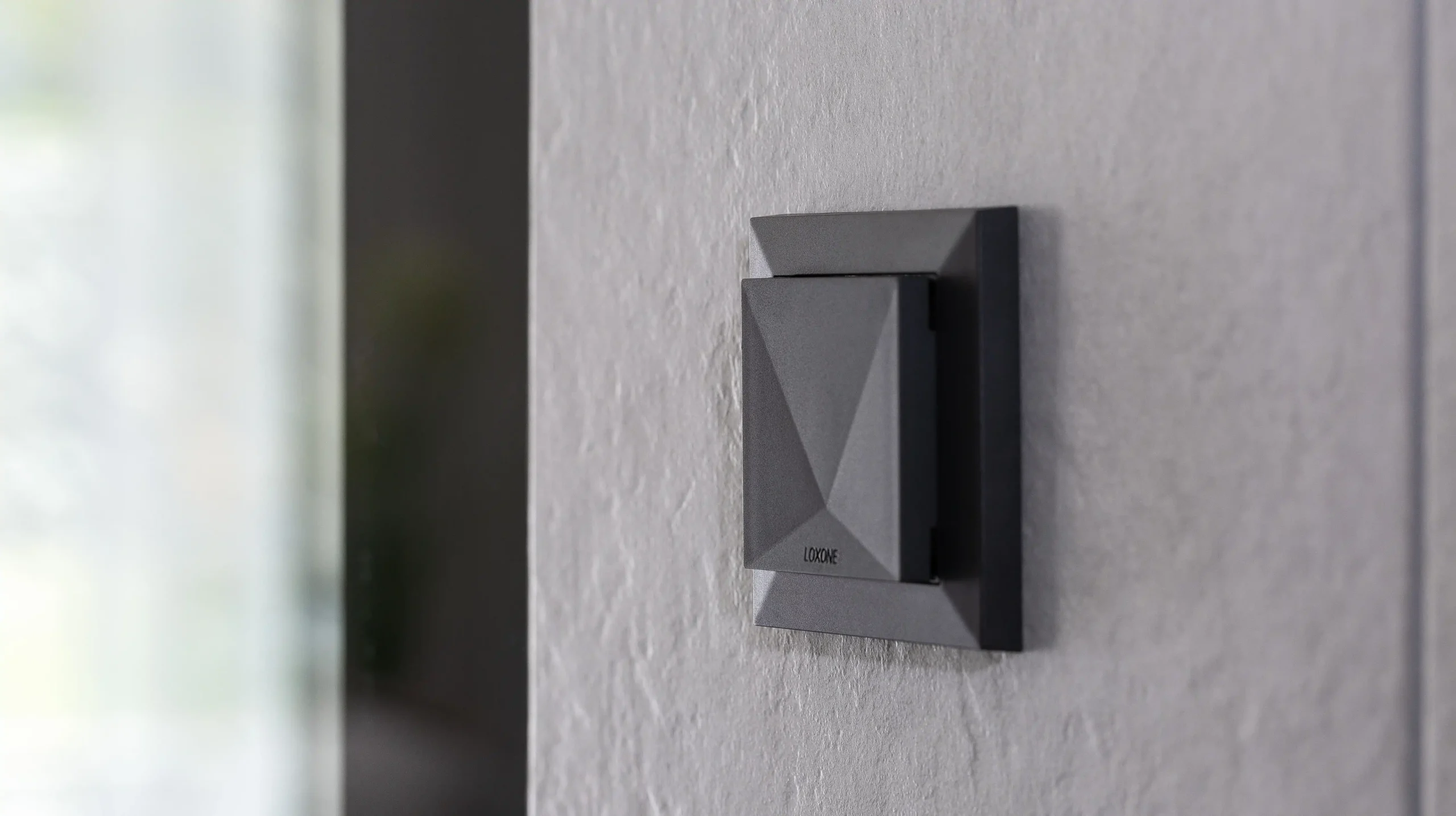 Een moderne, zwarte Loxone ruimte voeler gemonteerd op een grijze, gestructureerde muur. De sensor heeft een strak, geometrisch ontwerp met een subtiel reliëf en een matte afwerking. Op de achtergrond is een glazen oppervlak zichtbaar, wat duidt op een moderne en minimalistische interieurstijl. De sensor is ontworpen voor het meten van temperatuur luchtvochtigheid en CO2.