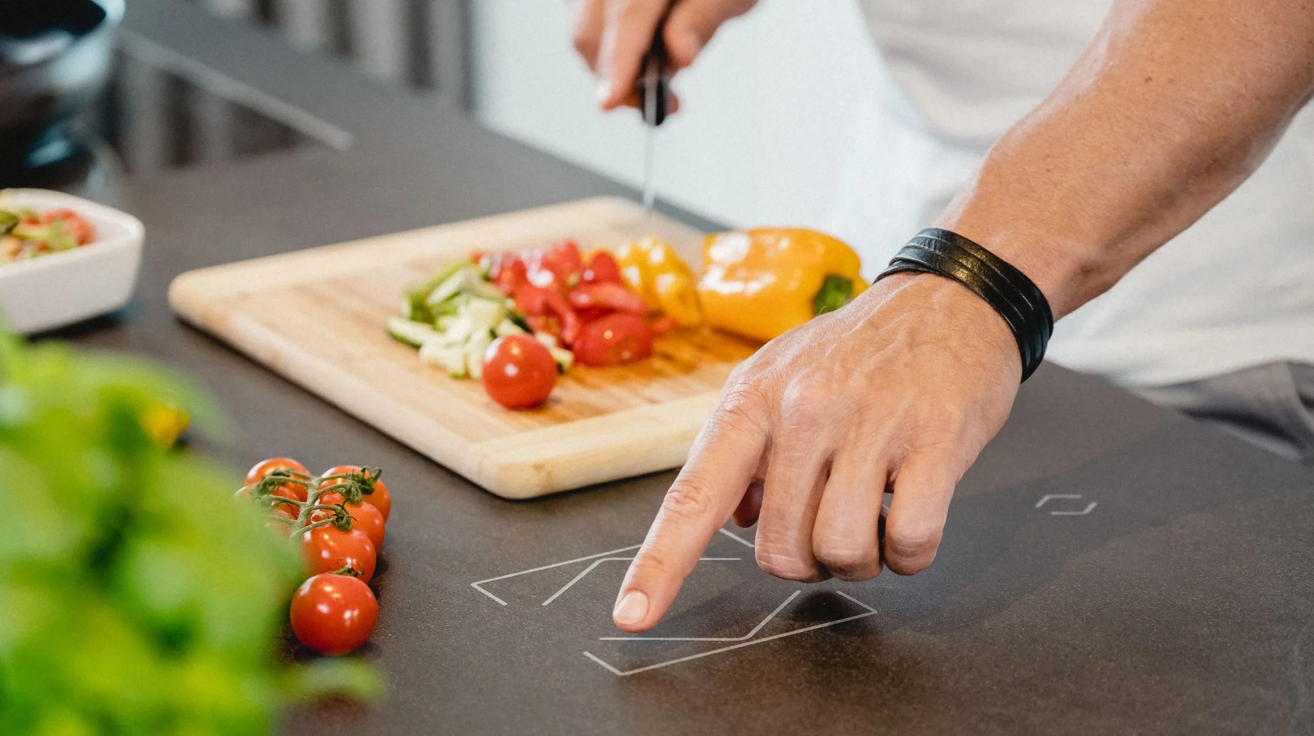 Een persoon snijdt groenten, waaronder cherrytomaten en een gele paprika, op een houten snijplank in een moderne keuken. Op de voorgrond ligt een tros tomaten naast een pot verse kruiden. De persoon bedient met één vinger een geïntegreerd Loxone touch surface op het donkere keukenblad, waarop bedieningssymbolen zichtbaar zijn. Dit suggereert het gebruik van slimme verlichting voor een efficiënte kookervaring.